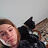 woman, cat, black_cat, pet, indoor, portrait, shoulder, collar, tag, clock, brick_wall, casual, expression, young_adult, plaid_shirt, face, feline, person, home, cute
