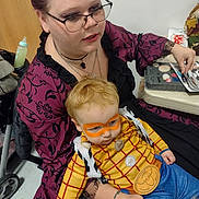 Niklous is registered to the contest to win money with this photo: woman, toddler, child, costume, face_paint, indoor, glasses, purple_dress, toy_story, woody_costume, orange_face_paint, holding, seated, table, makeup_kit, necklace, tattoo, playful, hair, person