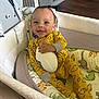 baby, infant, bassinet, yellow_onesie, cloud_pattern, football, toy, dinosaur_bedding, smiling, happy, indoor, cozy, child, cute, sleepwear, resting, crib, furniture, person, portrait
