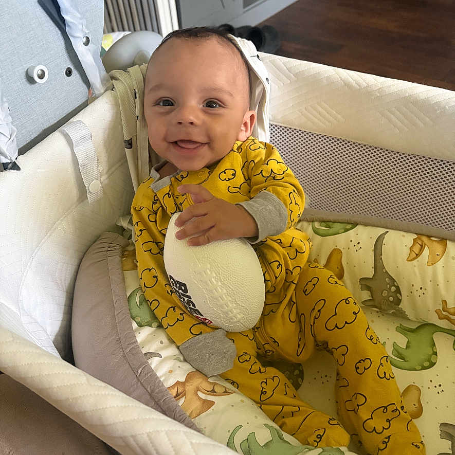 Shilo is registered to the contest to win money with this photo: baby, bassinet, child, cloud_pattern, cozy, crib, cute, dinosaur_bedding, football, furniture, happy, indoor, infant, person, portrait, resting, sleepwear, smiling, toy, yellow_onesie