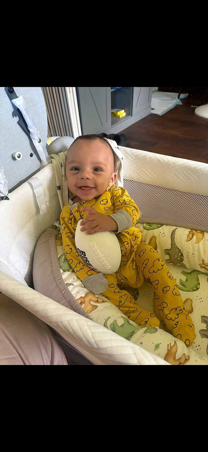 Shilo is registered to the contest to win money with this photo: baby, infant, bassinet, yellow_onesie, cloud_pattern, football, toy, dinosaur_bedding, smiling, happy, indoor, cozy, child, cute, sleepwear, resting, crib, furniture, person, portrait