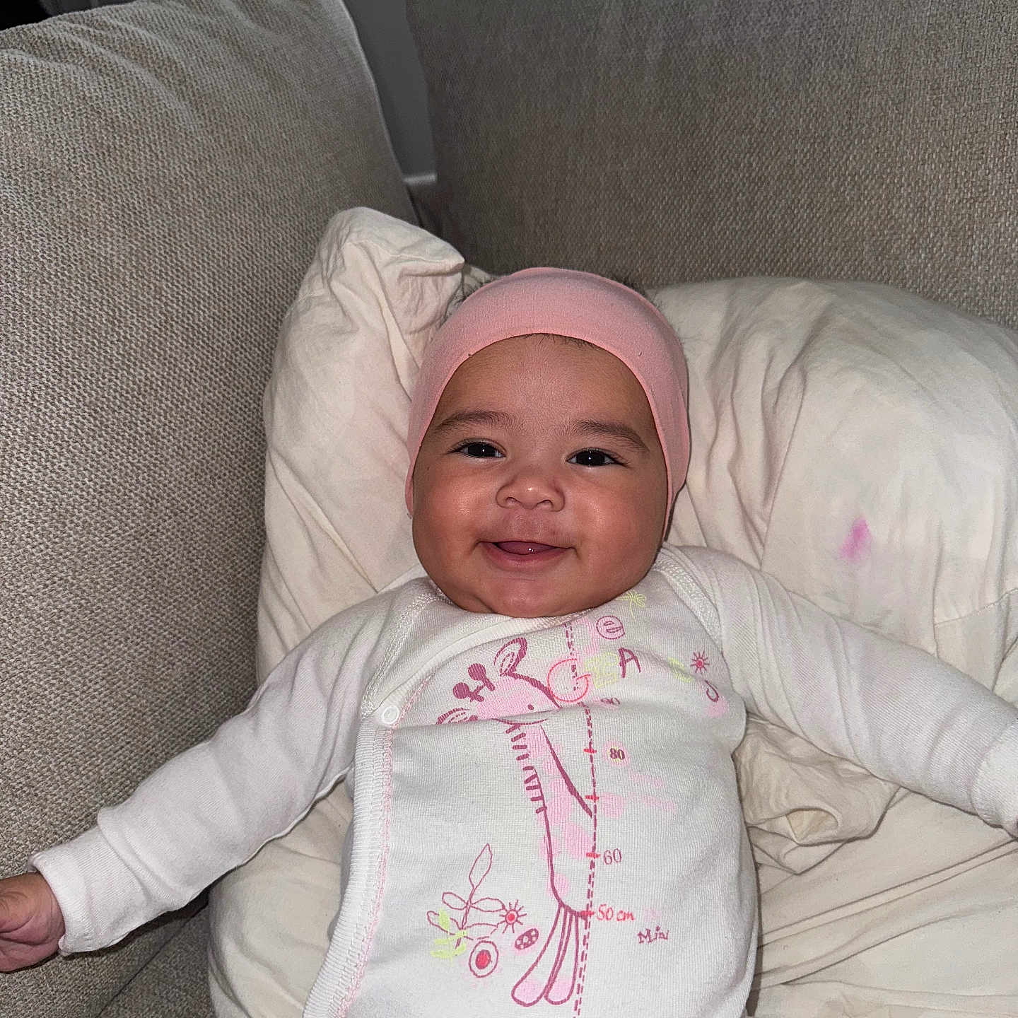 Aïtaynah participe au concours pour gagner de l'argent avec cette photo : baby, child, couch, cozy, cute, fabric, giraffe_design, happy, home, indoors, infant, person, pillow, pink_hat, portrait, relaxing, sitting, smiling, white_onesie, young_child
