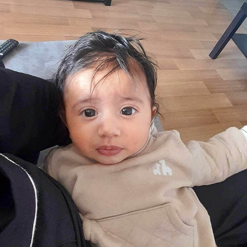 Nouh a rejoint le concours — aidez-le/la à gagner de superbes lots ! baby, child, indoor, person, face, clothing, sweater, pants, hair, living_room, floor, furniture, television, remote_control, sitting, holding, curious, young_child, home, casual