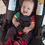 baby, car_seat, child, smiling, happy, infant, sneakers, colorful_clothing, straps, floor_tiles, red_shoes, cute, indoor, young_child, head, face, person, feet, jeans, laughter