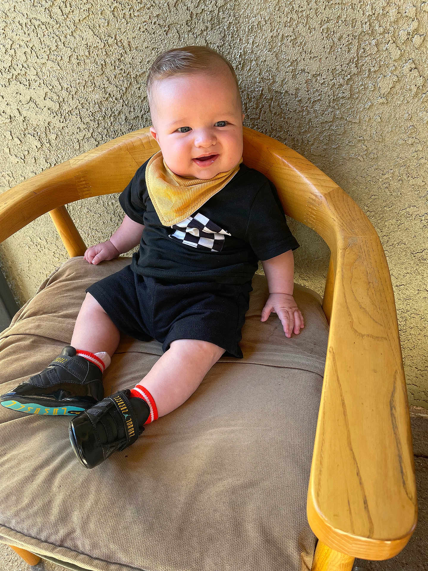 Xander Haze is registered to the contest to win money with this photo: baby, child, smile, black_clothing, yellow_bib, sitting, wooden_chair, cushion, shoe, sock, indoor, person, happy, cute, face, head, skin, texture, light, background