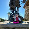 toddler, child, statue, horse, bronze_statue, pointing, outdoor, sunny, blue_sky, building, stone_platform, pink_shoes, casual_clothing, daytime, public_place, water_tower, sculpture, person, happy, smiling