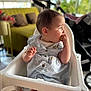 baby, blurred_background, child, clothing, couch, curious, cute, daylight, eating, furniture, high_chair, home, indoor, infant, person, pillows, seated, short_sleeves, snack, stroller