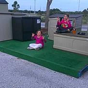 Brandon is registered to the contest to win money with this photo: calm, casual, child, cross_legged, daytime, fun, grass, gravel, green_platform, meditation_pose, nature, outdoor, pink_clothing, play, relaxation, sitting, sky, storage_shed, tree, two_children