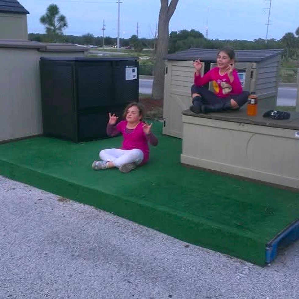 Brandon is registered to the contest to win money with this photo: calm, casual, child, cross_legged, daytime, fun, grass, gravel, green_platform, meditation_pose, nature, outdoor, pink_clothing, play, relaxation, sitting, sky, storage_shed, tree, two_children