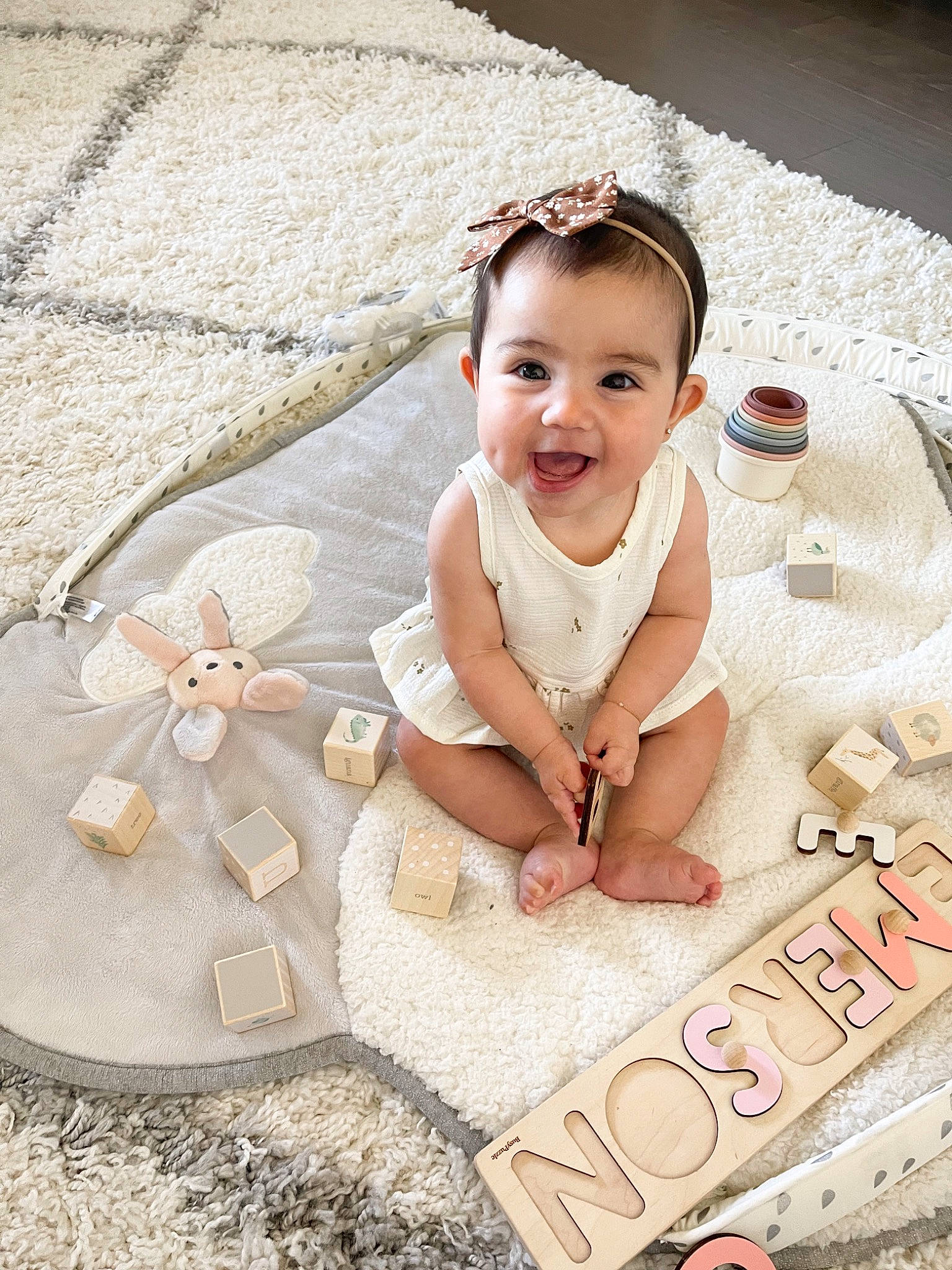 Emerson is registered to the contest to win money with this photo: baby, baby_toddler_clothing, child, dress, embellishment, facial_expression, fashion_accessory, flooring, fun, hair_accessory, happy, headpiece, person, pink, sand, sitting, skin, smile, toddler, white
