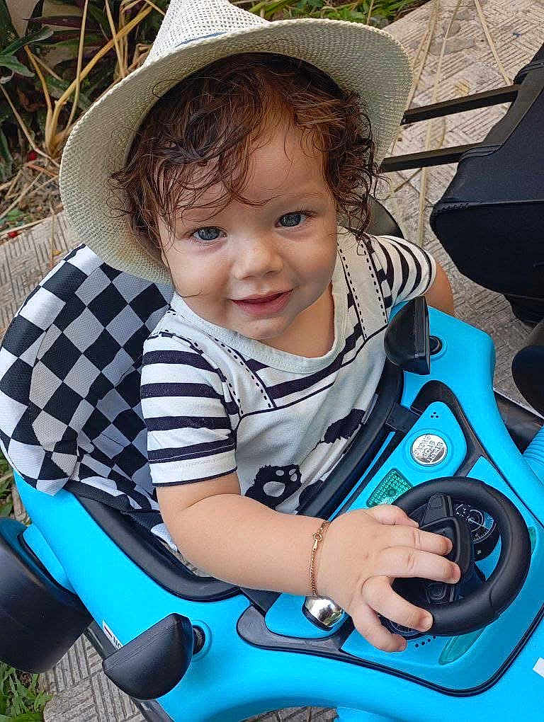 Demis joined the competition — help win amazing prizes! child, toddler, toy_car, hat, curly_hair, smiling, striped_shirt, bracelet, outdoor, steering_wheel, blue, seat, play, cute, person, happy, vehicle, young_child, fun, daytime