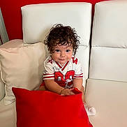 Demis joined the competition — help win amazing prizes! toddler, child, curly_hair, blue_eyes, white_couch, red_pillow, red_wall, smile, sitting, indoor, clothing, playful, person, cushion, bracelet, sock, portrait, cute, happy, home