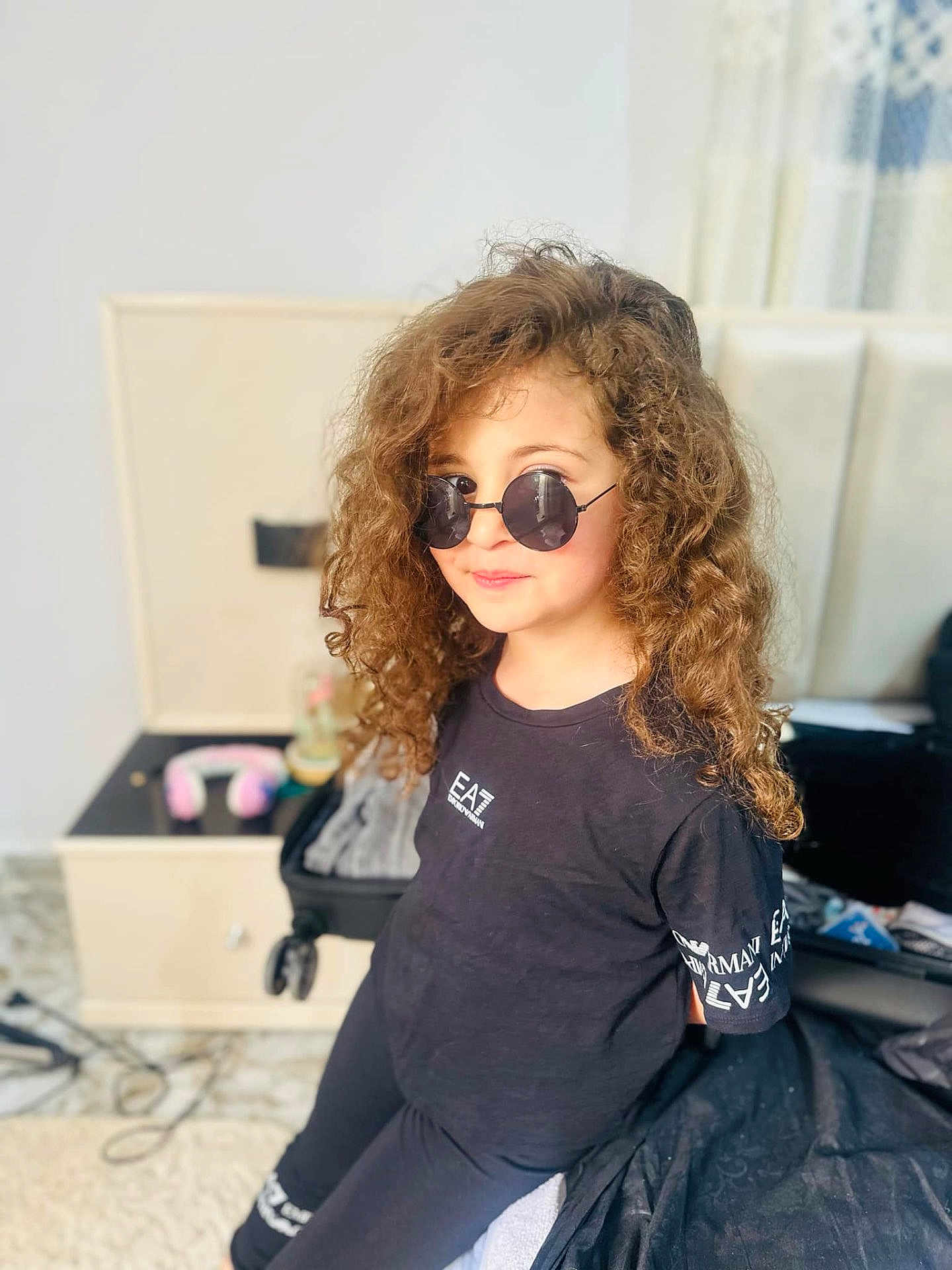Hadjira participe au concours pour gagner de l'argent avec cette photo : child, curly_hair, sunglasses, black_clothing, indoor, bedroom, nightstand, headphones, suitcase, person, casual, fashion, portrait, young, smile, curly, hair, cool, relaxed, furniture