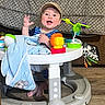 baby, blanket, blue_clothing, cap, carpet, child, cute, drool, floor, furniture, happy, indoor, infant, person, playful, seated, smiling, toy, walker, waving