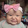 baby, child, face, headband, pink_bow, expression, indoor, blurred_background, clothing, cute, infant, smile, scrunched_face, chubby_cheeks, person, portrait, young_child, apparel, headwear, happy