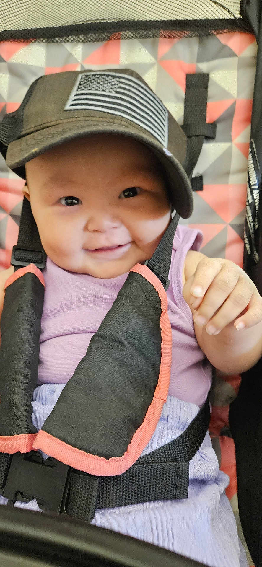 Valerian Jo is registered to the contest to win money with this photo: baby, smiling, cap, american_flag, stroller, child, face, hand, purple_clothing, safety_harness, patterned_background, infant, cute, happy, portrait, closeup, seatbelt, child_seat, baby_girl, playful