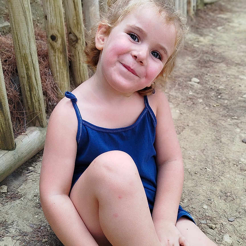 Lu-Ann a rejoint le concours — aidez-le/la à gagner de superbes lots ! blue_dress, casual, child, curly_hair, dirt_path, footwear, girl, happy, nature, outdoor, person, playful, portrait, sandals, sitting, skin, smiling, sunlight, wooden_fence, young