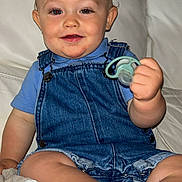 Kéylan a rejoint le concours — aidez-le/la à gagner de superbes lots ! baby, blanket, blue_shirt, child, clothing, couch, curly_hair, denim_overalls, face, hand, happy, indoor, pacifier, person, sitting, skin, smiling, sock, toddler, white_couch