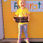 Emalee joined the competition — help win amazing prizes! child, clothing, congratulations_sign, door, front_door, girl, hair_clip, happy, holding, leggings, mat, outdoor, person, preschool, sign, smiling, sneakers, standing, sunlight, yellow_door
