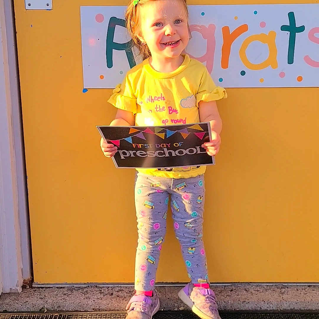 Emalee joined the competition — help win amazing prizes! child, clothing, congratulations_sign, door, front_door, girl, hair_clip, happy, holding, leggings, mat, outdoor, person, preschool, sign, smiling, sneakers, standing, sunlight, yellow_door
