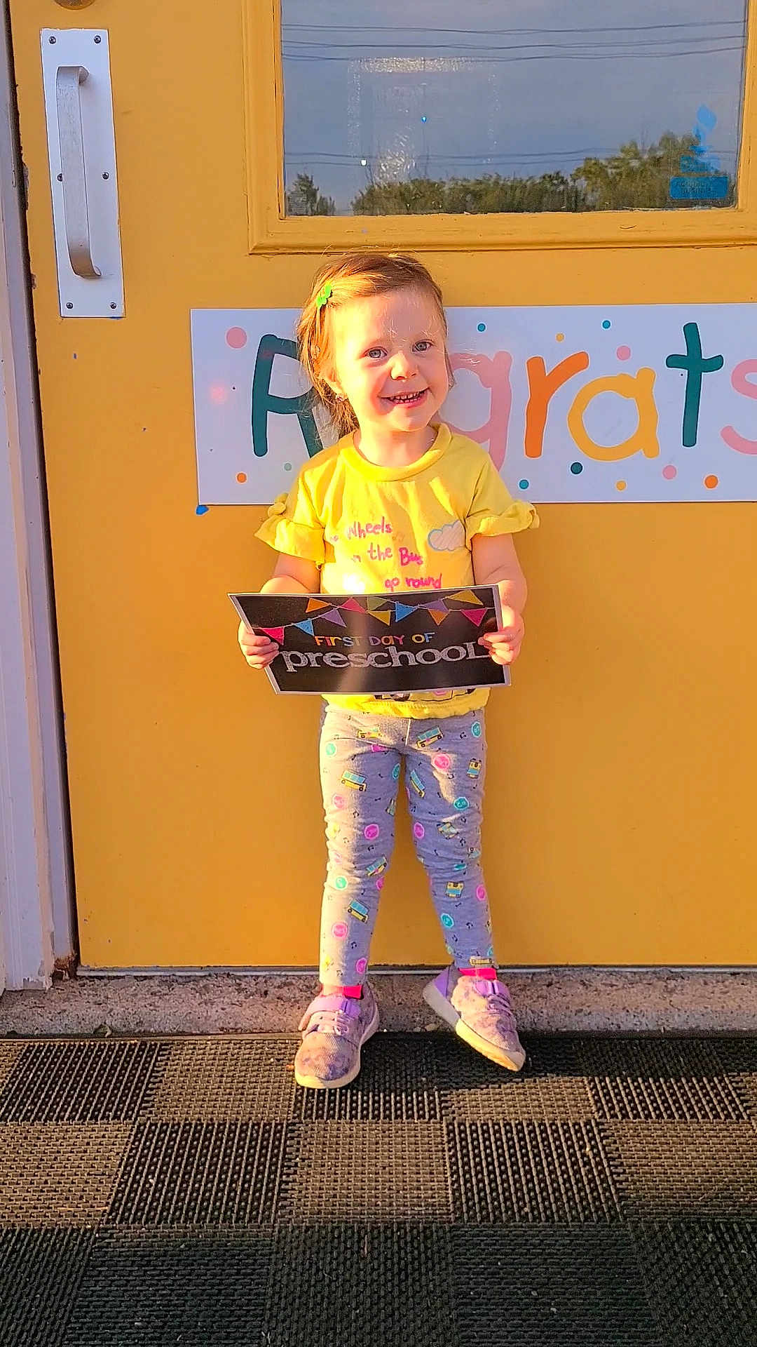 Emalee joined the competition — help win amazing prizes! child, clothing, congratulations_sign, door, front_door, girl, hair_clip, happy, holding, leggings, mat, outdoor, person, preschool, sign, smiling, sneakers, standing, sunlight, yellow_door