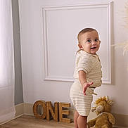 Oliver is registered to the contest to win money with this photo: baby, child, knitted_clothing, standing, smiling, plush_toy, wooden_letters, one, floor, wall, home_interior, curtains, barefoot, portrait, cute, happy, indoor, young_child, person, toddler