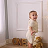 baby, child, knitted_clothing, standing, smiling, plush_toy, wooden_letters, one, floor, wall, home_interior, curtains, barefoot, portrait, cute, happy, indoor, young_child, person, toddler