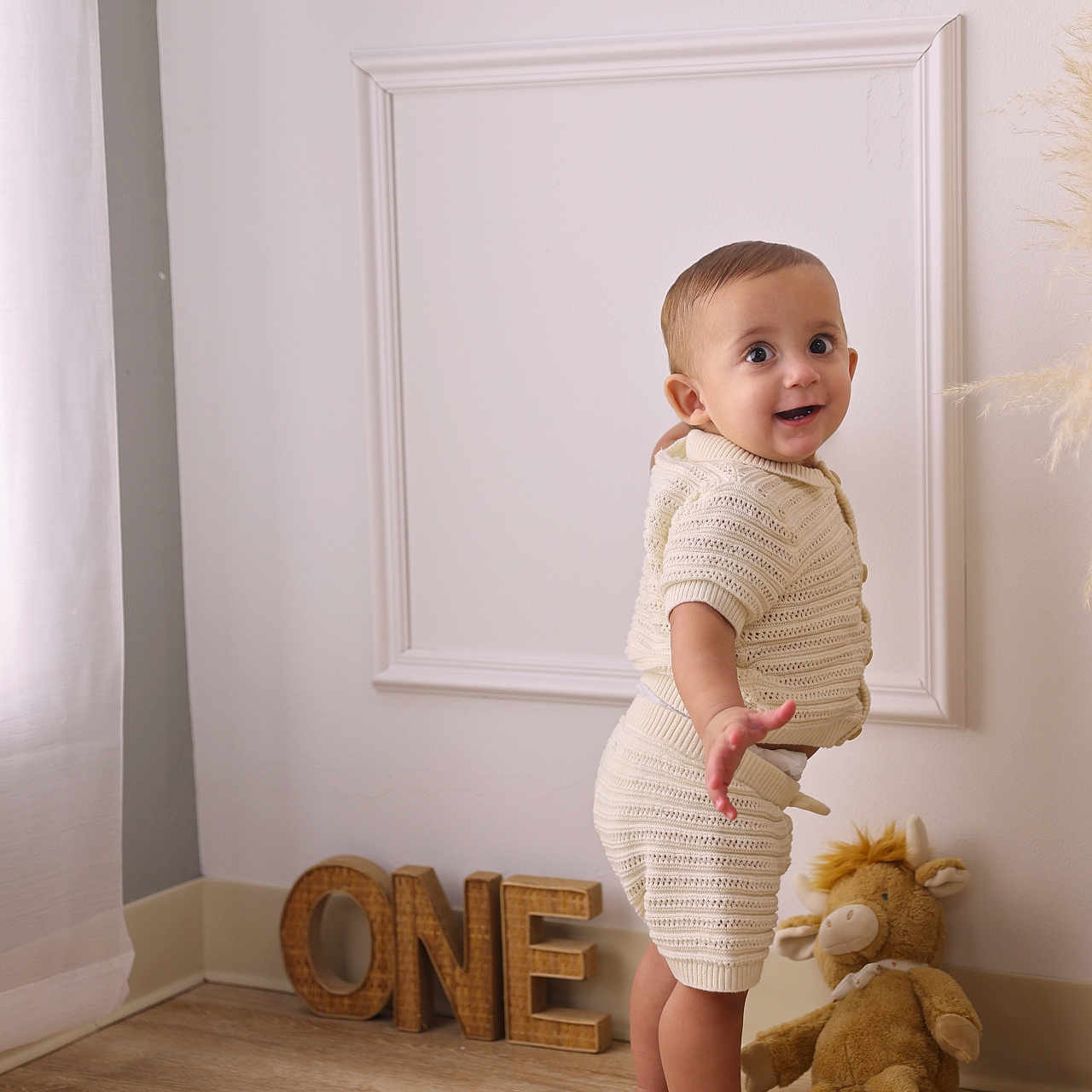 Oliver is registered to the contest to win money with this photo: baby, child, knitted_clothing, standing, smiling, plush_toy, wooden_letters, one, floor, wall, home_interior, curtains, barefoot, portrait, cute, happy, indoor, young_child, person, toddler