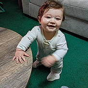 Oliver is registered to the contest to win money with this photo: baby, child, smile, onesie, standing, table, wood, carpet, green_carpet, couch, striped_couch, indoor, face, hand, hair, bottle, floor, happy, person, home