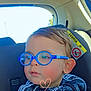 child, toddler, glasses, blue_glasses, eyewear, car_seat, car_interior, car_backseat, window, rear_window, seat_headrest, patterned_jacket, hoodie, heart_doodle, rosy_cheeks, ear, portrait, closeup, sunlight, safety_label
