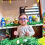 child, toddler, eyeglasses, blue_glasses, easter, easter_egg, easter_basket, chocolate_bunny, foil_wrapping, decorative_grass, green_grass, smiling, portrait, indoor, shelf, toys, plush_chick, painted_egg, cardboard_box, festive