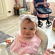 Joanie-May is registered to the contest to win money with this photo: baby, bow, child, clothing, curtains, face, floor, food_stains, hand, headband, high_chair, indoor, person, pink_bib, seat, smiling, stroller, table, toys, window