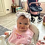 baby, bow, child, clothing, curtains, face, floor, food_stains, hand, headband, high_chair, indoor, person, pink_bib, seat, smiling, stroller, table, toys, window