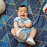 baby, infant, child, smiling, sitting, portrait, indoors, bed, bedspread, blue, geometric_pattern, clothing, light_blue_outfit, shoes, white_shoes, toy, pillow, happy, playful, photography