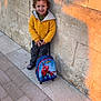 child, curly_hair, yellow_jacket, backpack, spiderman, stone_wall, sidewalk, smiling, outdoor, sunlight, shadow, happy, casual_clothing, young_boy, pavement, urban, portrait, standing, toy, daylight