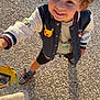 child, toddler, curly_hair, smiling, pokemon, jacket, outdoor, sunlight, shoes, shorts, happy, person, fashion, casual, standing, looking_up, playful, ground, shadow, daytime