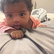 Reina is registered to the contest to win money with this photo: baby, infant, curly_hair, big_eyes, tongue_out, pink_onesie, hand, blanket, bed, close_up, portrait, indoor, cozy, soft_texture, adorable, crawling_pose, foreground, curious_expression, nursery_background, bedroom