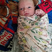 Everly joined the competition — help win amazing prizes! baby, infant, swaddled, blanket, floral_pattern, quilt, sports_theme, face, wide_eyes, fist, bed, bedding, colorful, curious, lying_down, indoors, soft_texture, closeup, person, newborn