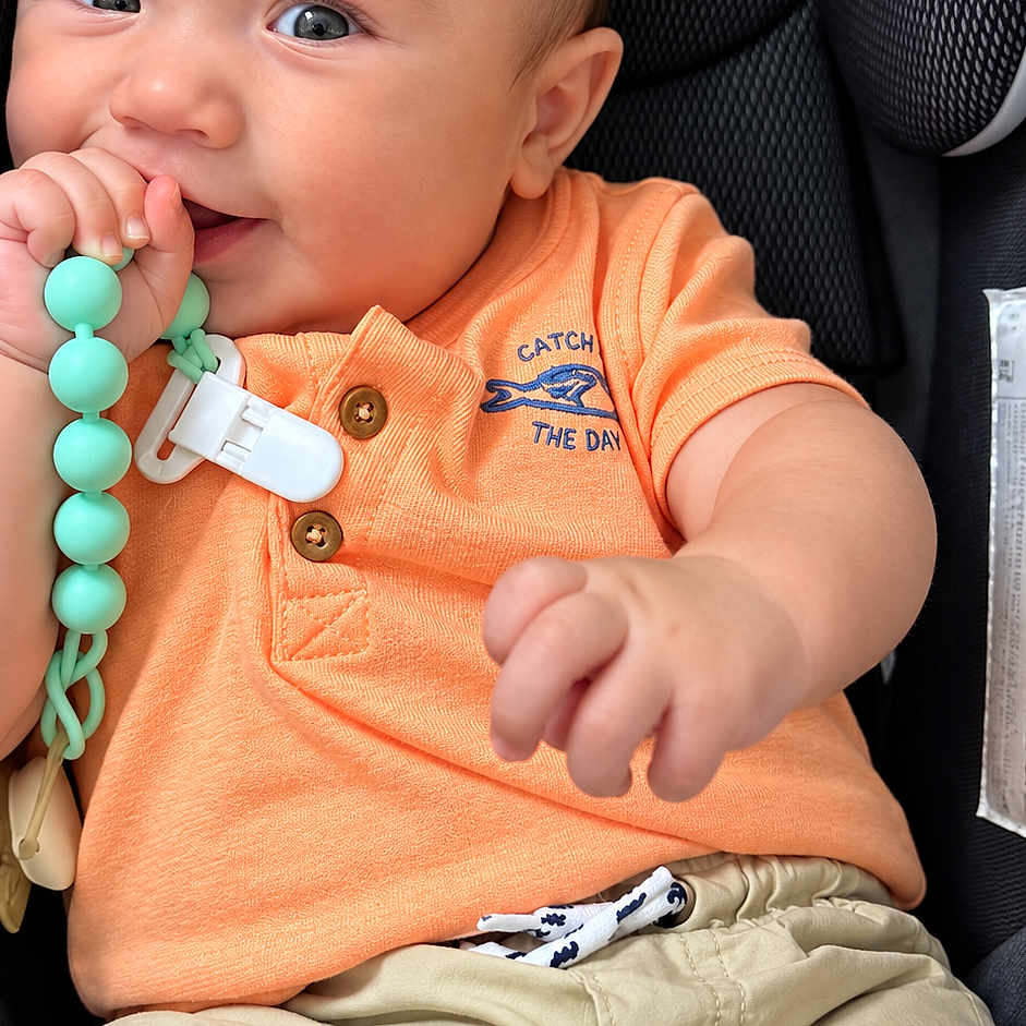 Dylan is registered to the contest to win money with this photo: baby, child, infant, car_seat, portrait, smiling, sitting, teething_toy, pacifier_clip, orange_shirt, beige_shorts, brown_hair, blue_eyes, curious, indoors, close_up, face, hands, casual_clothing, photography