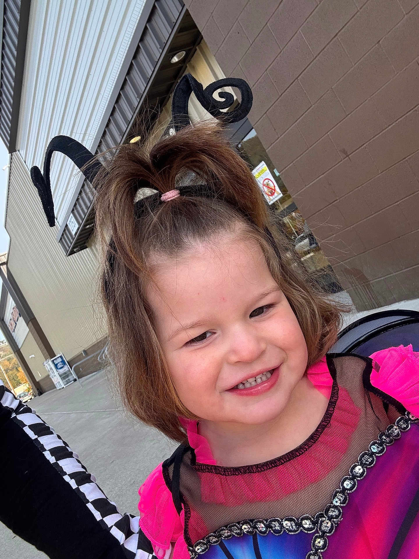 Brexley is registered to the contest to win money with this photo: child, toddler, costume, smile, headband, antennae, outdoor, building, sidewalk, brick_wall, happy, face, hair, pink, black, blue, clothing, window, daylight, person
