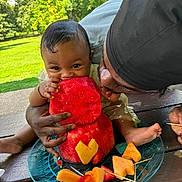 Naiari joined the competition — help win amazing prizes! baby, child, watermelon, fruit, outdoor, picnic, plate, table, greenery, person, adult, eating, summer, happy, smile, hand, foot, nature, food, family