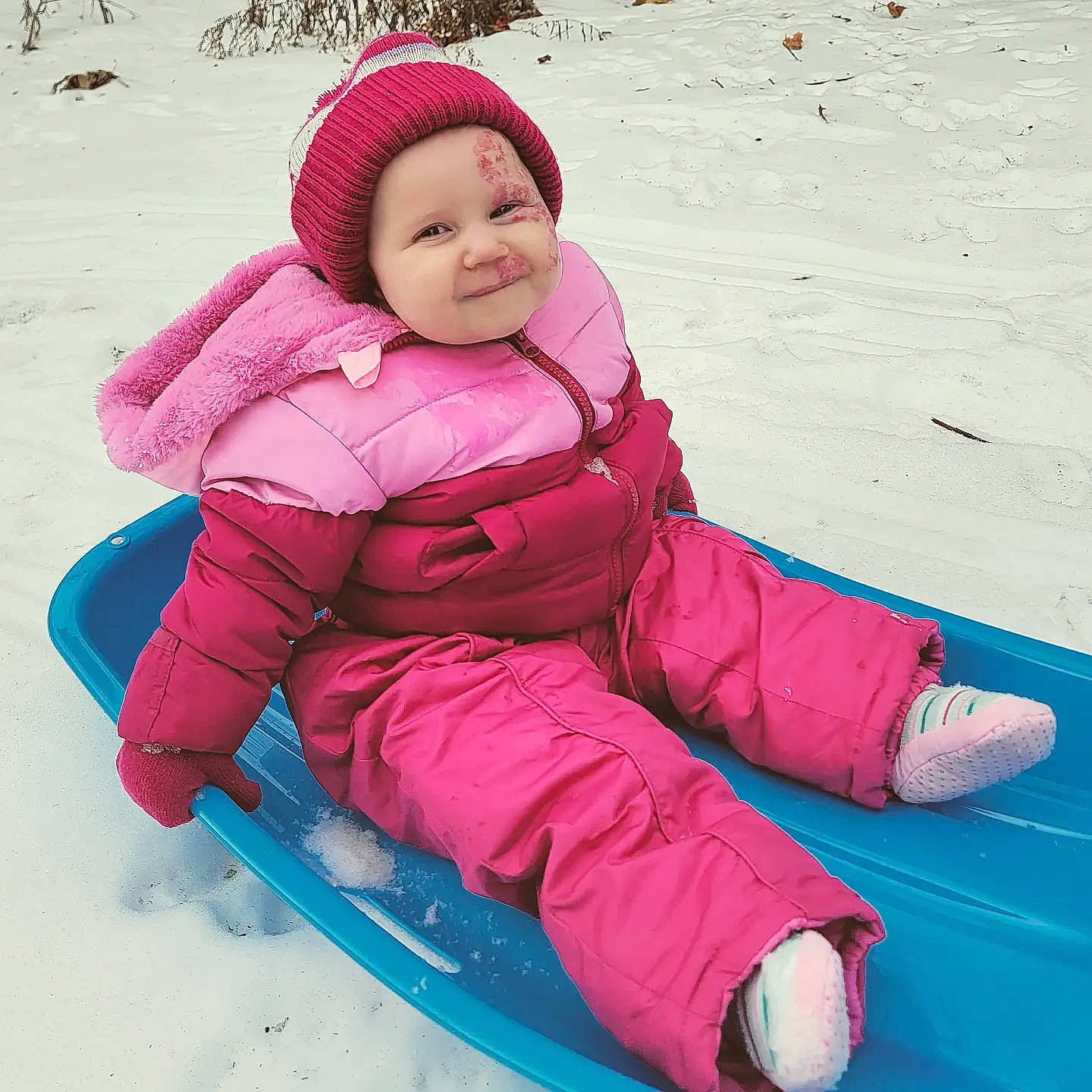 Charlie is registered to the contest to win money with this photo: baby_toddler_clothing, comfort, face, freezing, happy, headwear, human_body, jacket, joy, leisure, magenta, outdoor_recreation, outerwear, person, pink, recreation, sled, sledding, sleeve, smile
