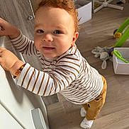 Elyo a rejoint le concours — aidez-le/la à gagner de superbes lots ! toddler, child, curly_hair, striped_shirt, brown_pants, socks, bracelet, drawer, wooden_floor, toy, box, indoor, standing, smile, face, person, home, radiator, flooring, playtime