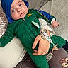 Noah participe au concours pour gagner de l'argent avec cette photo : baby, child, blue_hat, green_outfit, pacifier_clip, hand, adult, cushion, cartoon_print, pants, indoor, curious_look, wide_eyes, soft_texture, casual_clothing, seated, comfort, closeup, person, holding