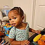 toddler, child, toy_food, play_table, braided_hair, curious_expression, floral_shirt, indoor, carpet, chair, bucket, stuffed_animals, doll, door, hand, face, person, playing, room, home