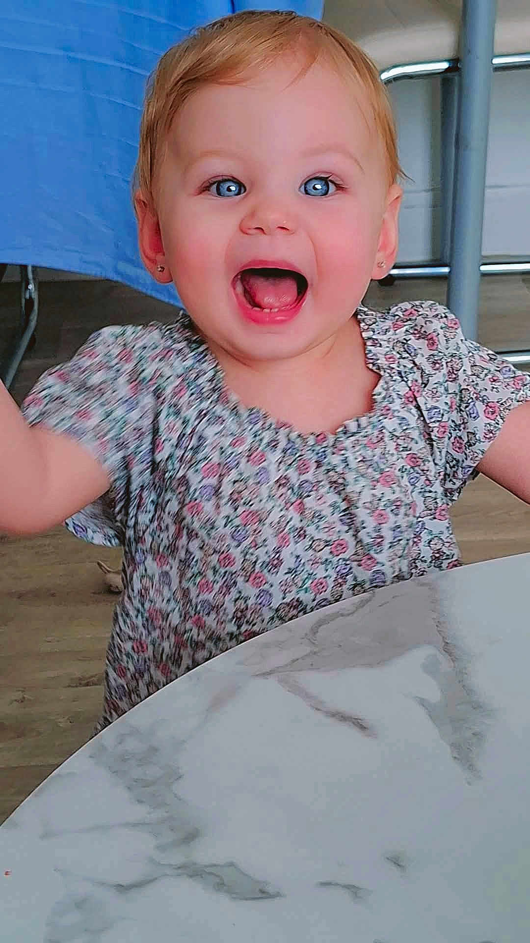 énaya participe au concours pour gagner de l'argent avec cette photo : toddler, child, happy, smiling, blue_eyes, floral_dress, table, marble_table, indoor, person, face, arms_raised, joy, young_child, cute, earrings, wooden_floor, chair, background, expression