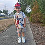 child, boy, spiderman, cap, shorts, sneakers, sidewalk, outdoor, trees, grass, residential_area, smile, pose, street, daylight, casual_clothing, person, nature, happy, summer