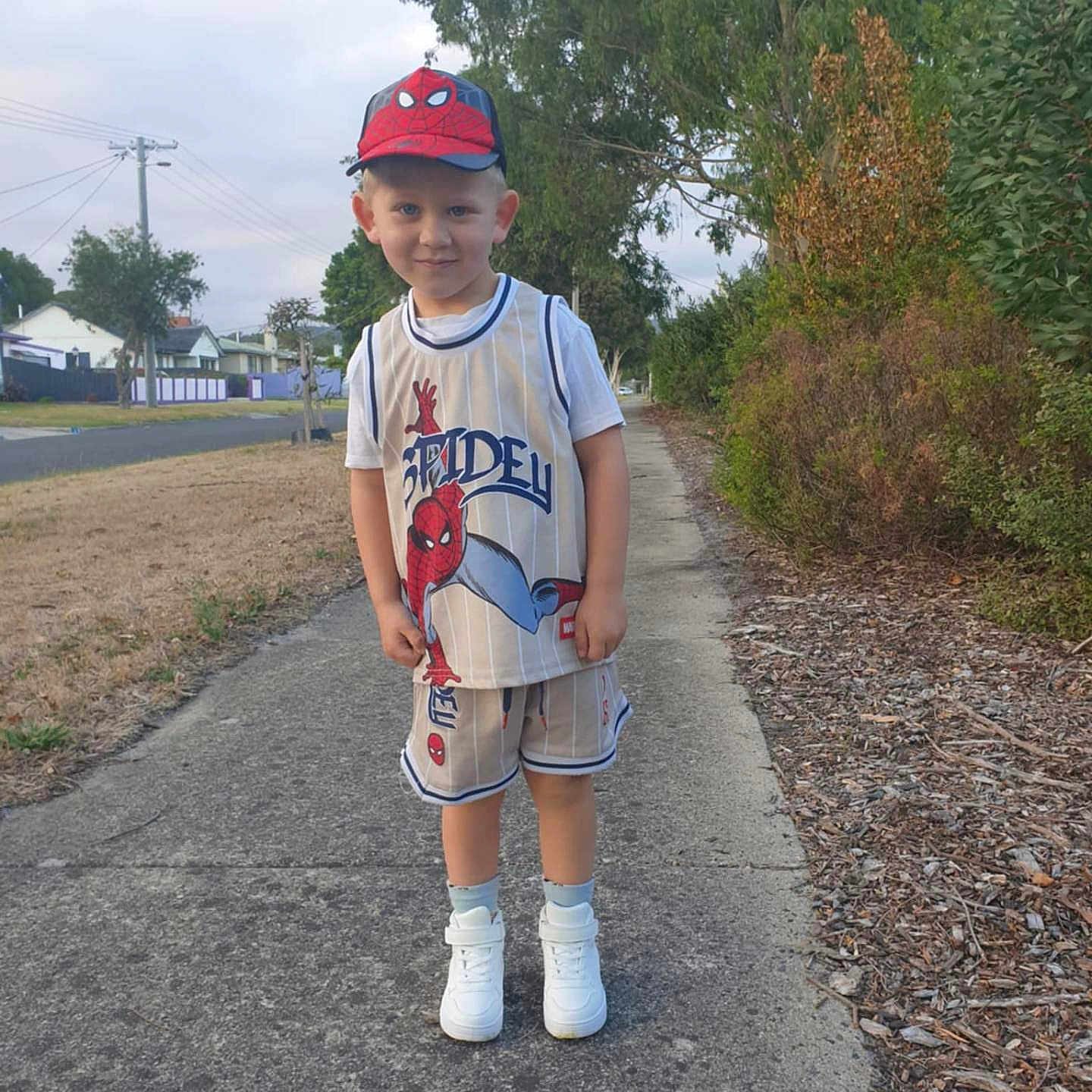 Cruze is registered to the contest to win money with this photo: boy, cap, casual_clothing, child, daylight, grass, happy, nature, outdoor, person, pose, residential_area, shorts, sidewalk, smile, sneakers, spiderman, street, summer, trees