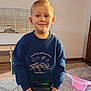 child, boy, indoor, smiling, blue_sweatshirt, jeans, toy, pin_art, pet_cage, carpet, table, wooden_floor, blonde_hair, face, person, home, hat, fuzzy, kindergarten, casual_clothing