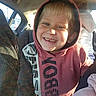 child, boy, hoodie, smile, car_interior, sunlight, person, curly_hair, jacket, seat, window, daylight, happy, portrait, casual_clothing, seatbelt, vehicle, young_child, warm_light, candid